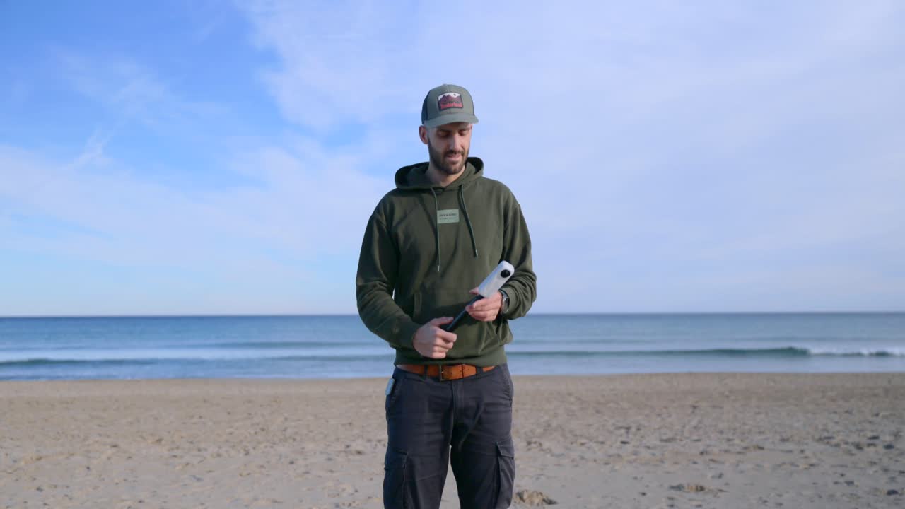 Un uomo realizza un vlog con una fotocamera a 360 gradi su un selfie stick. Si trova su una spiaggia sabbiosa con l'oceano sullo sfondo. Ideale per concetti di viaggio, tecnologia e creazione di contenuti.