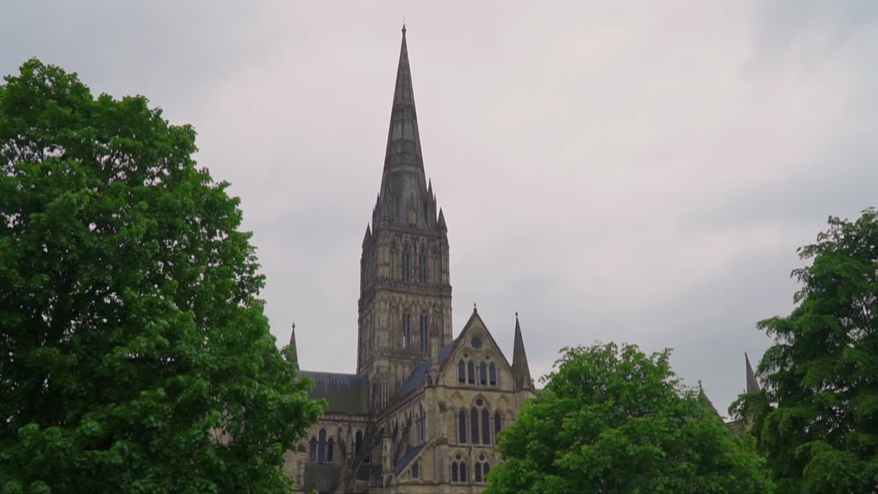 Iconic spire of Salisbury Cathedral rises behind vibrant summer foliage in Wiltshire, England, showcasing Gothic architecture in a historic religious setting.