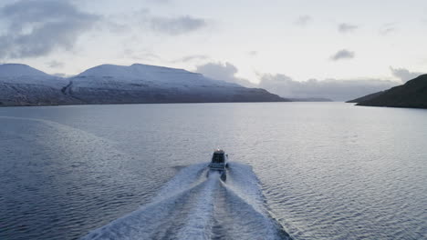 faroe islands, 4k aerial tracking behind fishing boat with snow covered mountains in the background