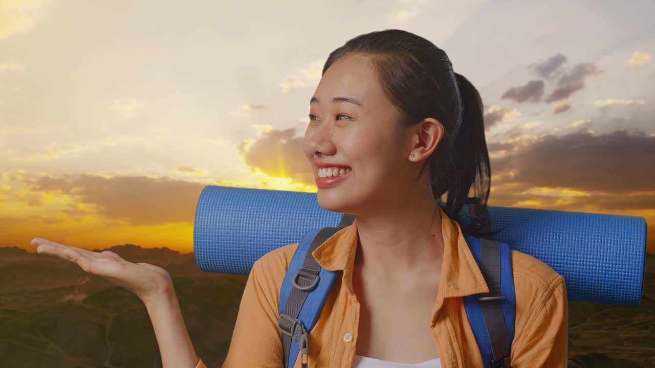 Close Up Of Asian Female Hiker With Mountaineering Backpack Smiling And Pointing To Side While Standing On The Top Of Mountain During Sunset Time