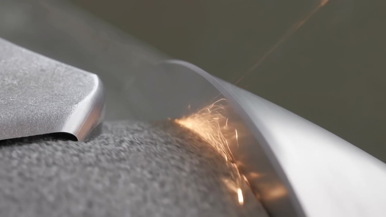 Makroansicht aus niedrigem Winkel zeigt zwei Metallkomponenten, die sich auf strukturiertem grauem Stoff zusammenpressen und orange Funken an der Naht erzeugen, mit Reibung, Hitze, Reflexion und präzisem industriellem Oberflächendetail.