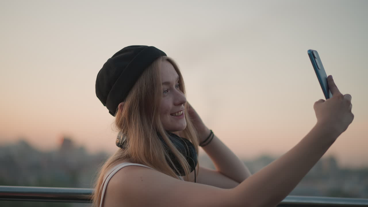 sunset rooftop selfie with headphones, young woman in beanie captures contemplative portrait with smartphone, soft golden light, blurred city skyline, gentle breeze, relaxed expression, casual style