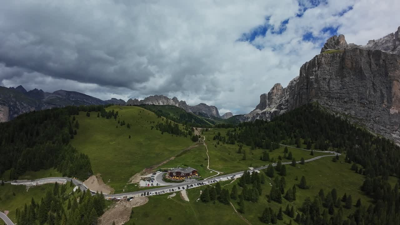 Luftfoto af Dolomitterne, Italien. Dal under overskyet himmel, tårnhøje kalkstensklipper, frodigt grønne enge, bugtet bjergvej, der fører til en lille alpelandsby, tæt fyrreskov, stemningsfuldt dramatisk lys