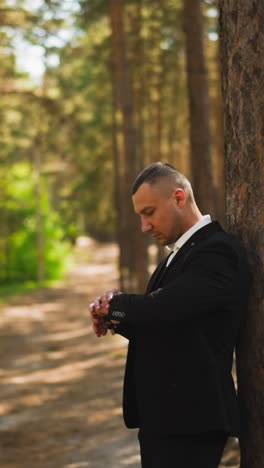 man wearing elegant black suit checks time waiting for beloved girlfriend near pine tree in shady coniferous forest side view slow motion
