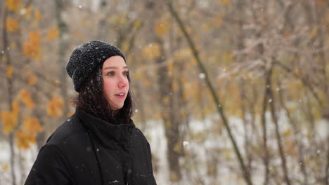 lady in black hoodie carrying guitar with hands in pockets walking through snow-covered forest path, surrounded by bare trees and falling snow, creating a serene winter scene