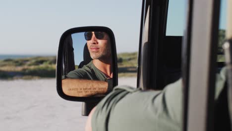 Un-Hombre-Caucásico-Feliz-Con-Gafas-De-Sol-Sentado-En-El-Coche-Reflejado-En-El-Espejo-Lateral-En-Un-Día-Soleado-En-La-Playa