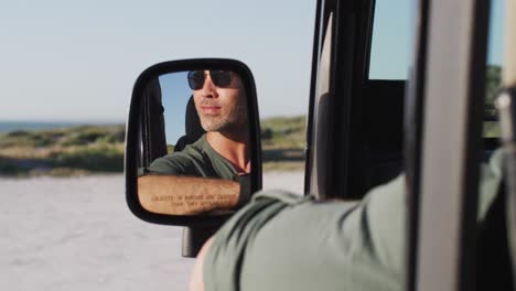 feliz hombre caucásico con gafas de sol sentado en el coche reflejado en el espejo lateral en un día soleado en la playa