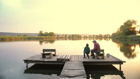 Rear-view-of-a-teen-boy-sitting-with-his-grandfather-on-the-lake-pier,-talking-and-fishing-together