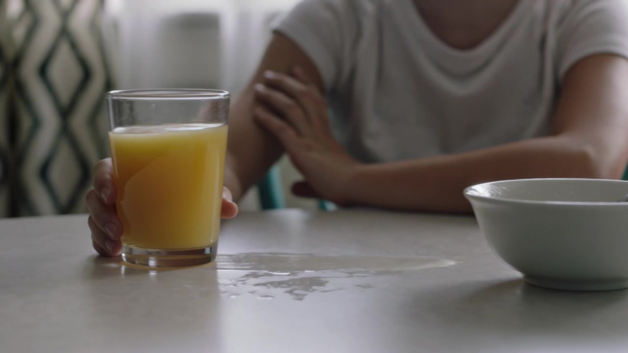 Mujer con camiseta extendiendo la mano y agarrando un vaso de zumo, moviéndose a la izquierda en la mesa de casa, colocándolo lejos de un derrame.