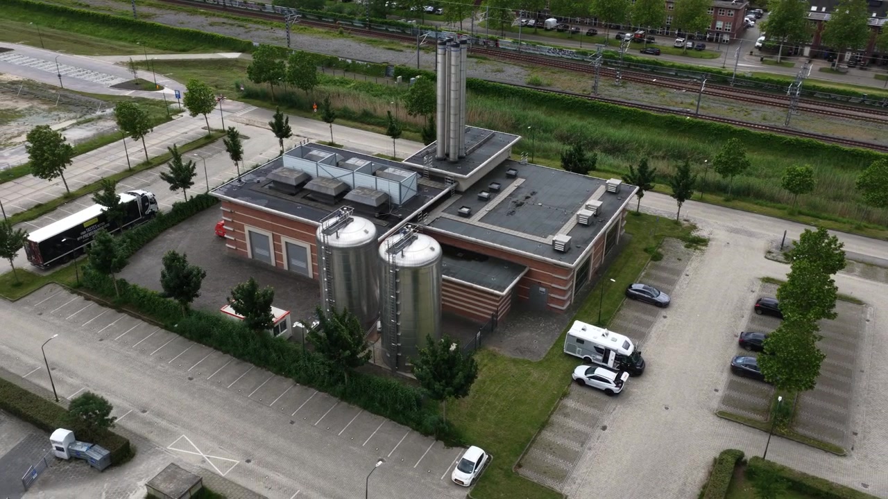 High angle rotating shot of a modern industrial facility featuring a tall chimney and large silver storage silos, situated next to parking lots and railroad tracks in a suburban area