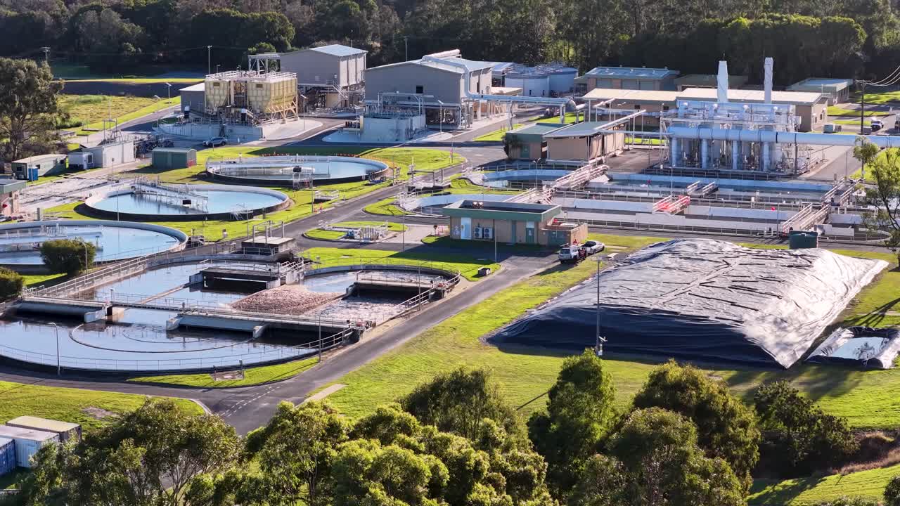 Aerial camera pans across a modern wastewater treatment plant surrounded by greenery, showing water tanks, buildings, and infrastructure in bright daylight