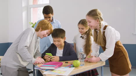 multiethnic students group around a desk where an student is sitting using a mobile in english classroom