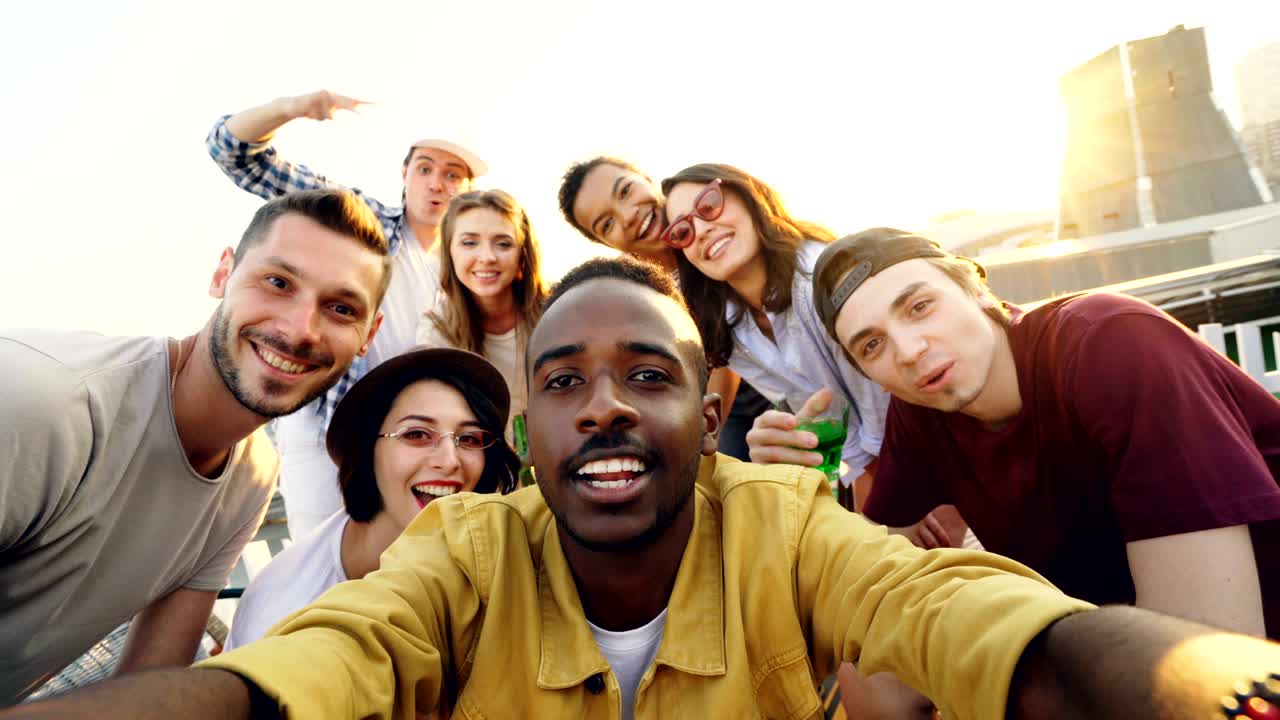 punto de vista de un apuesto hombre afroamericano tomando una selfie con sus amigos felices, la gente está mirando a la cámara, sonriendo y posando con botellas en una fiesta en el techo.
