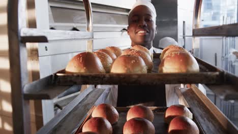 Happy-african-american-male-baker-in-bakery-kitchen,-holding-baking-sheet-with-rolls-in-slow-motion