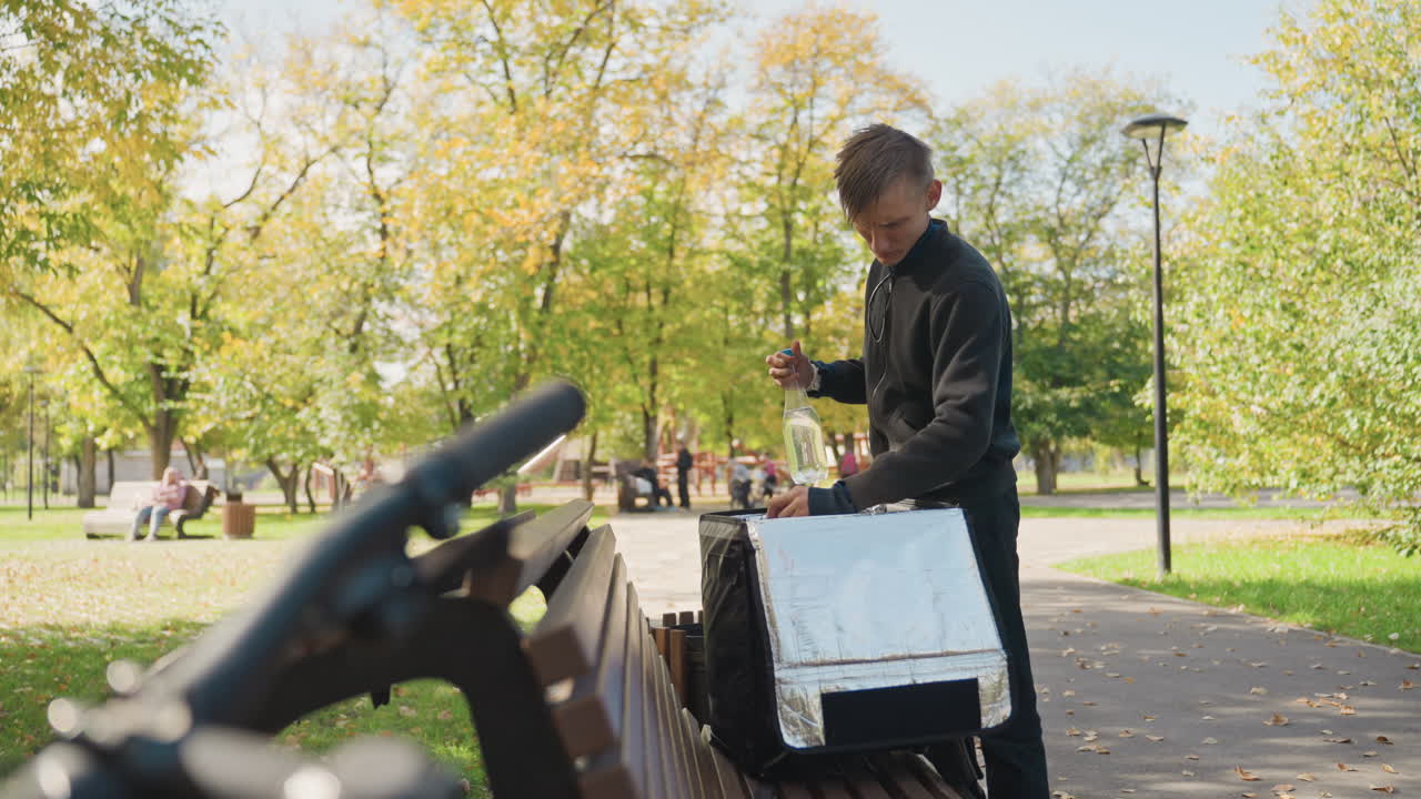 En kurér åbner en isoleret taske på en bænk, tager en flaske drik og tjekker ordren. Han tager en slurk, mens cyklen hviler i forgrunden. Efterårstræer og en lygtepæl indrammer den afslappede frokostpause i byen for leveringen