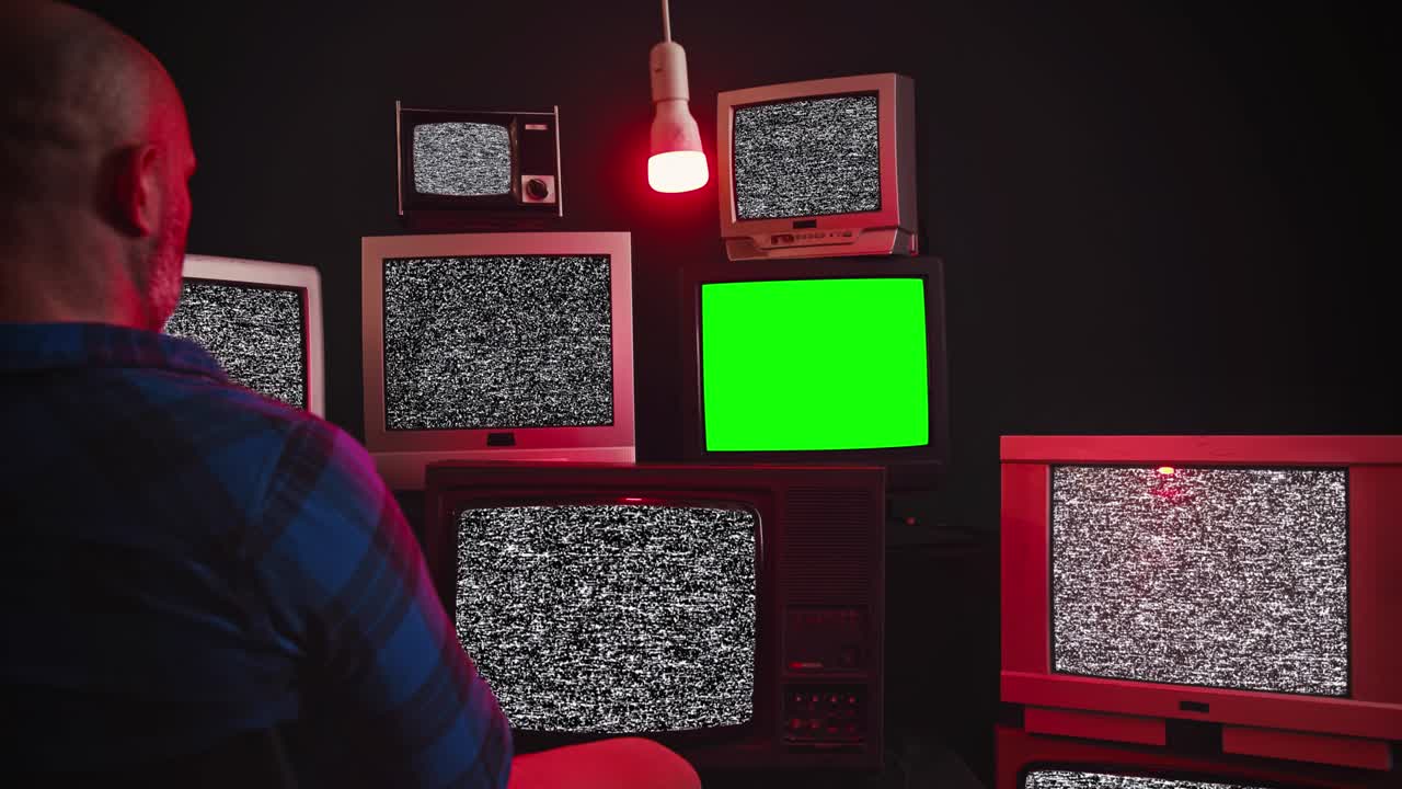A man sits in front of group of 9 vintage TV with chroma key screens in room with black wall