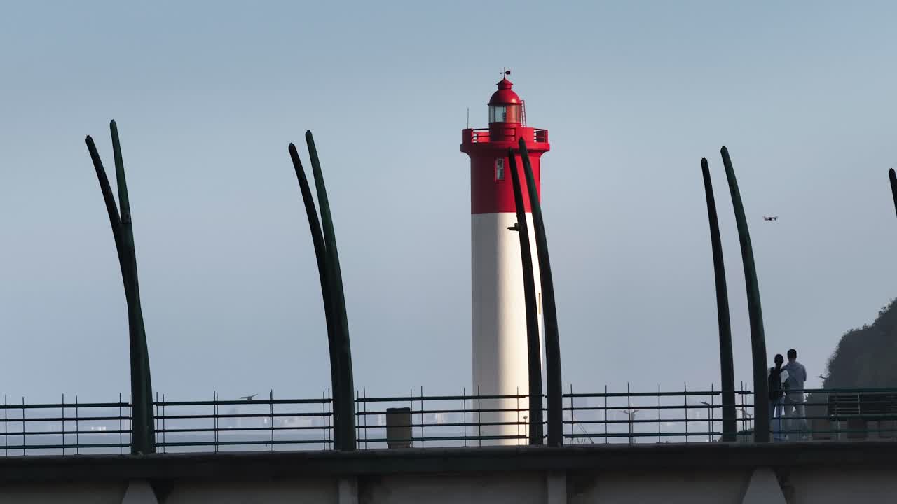 Red and white lighthouse at Umhlanga with silhouettes of a couple on the bridge, clear skies