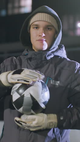 entrenador rodando una pelota de fútbol en sus manos mientras usa una sudadera con capucha y guantes en un campo deportivo urbano durante la práctica nocturna, edificios débilmente iluminados y luces bokeh