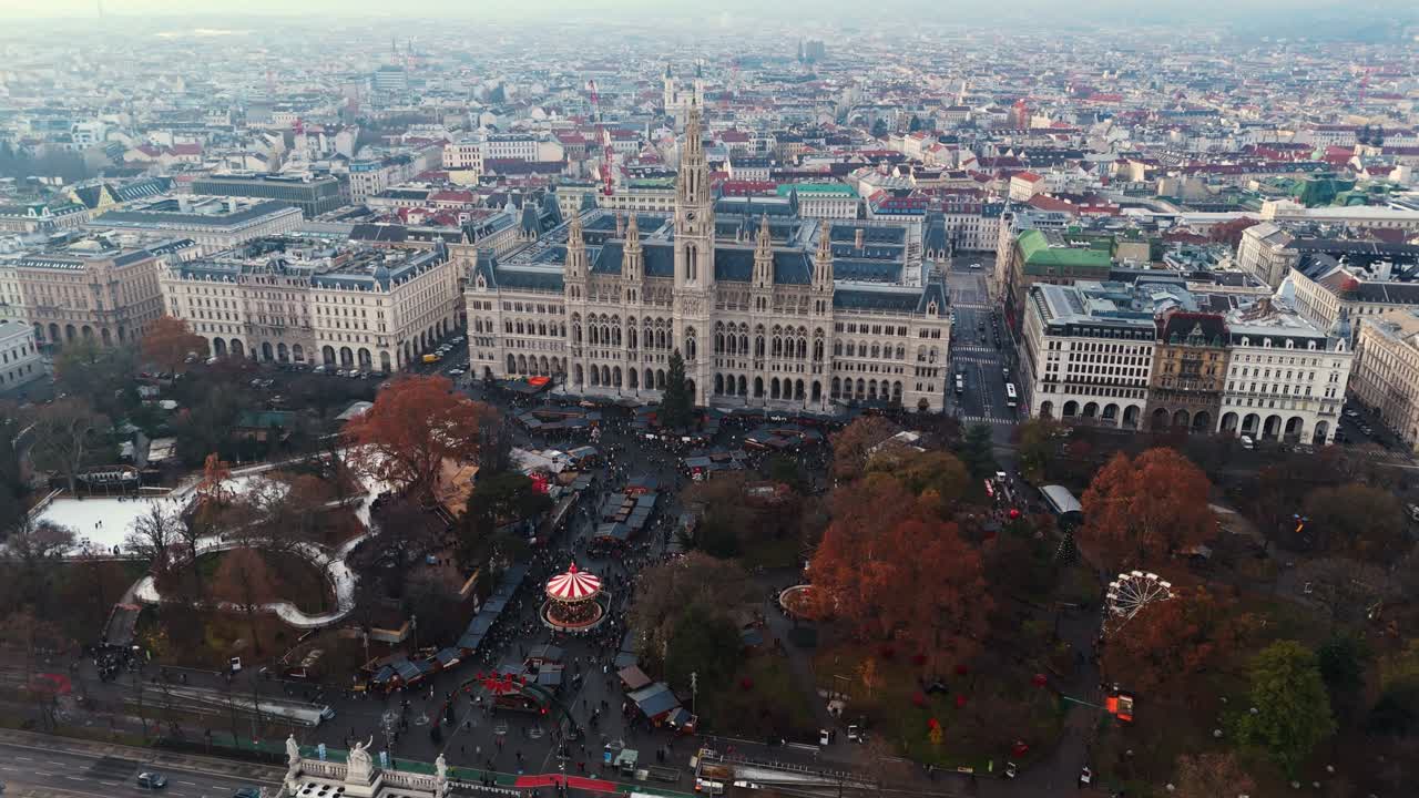 Ripresa aerea orizzontale con drone del mercatino di Natale di Rathausplatz a Vienna, che mostra file di bancarelle festive, fitte folle invernali sottostanti ed edifici storici che circondano la piazza