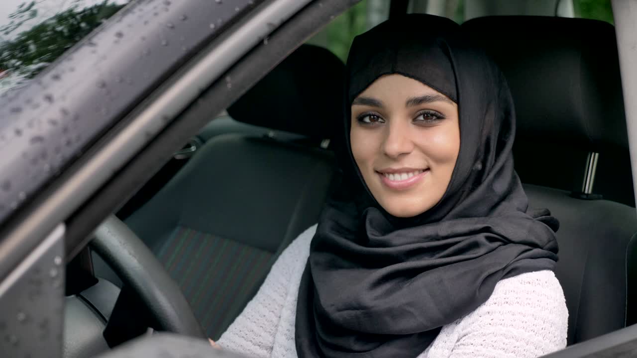 Young beautiful muslim woman in hijab is sitting in car in rainy weather, watching at camera, religiuos concept, transport concept