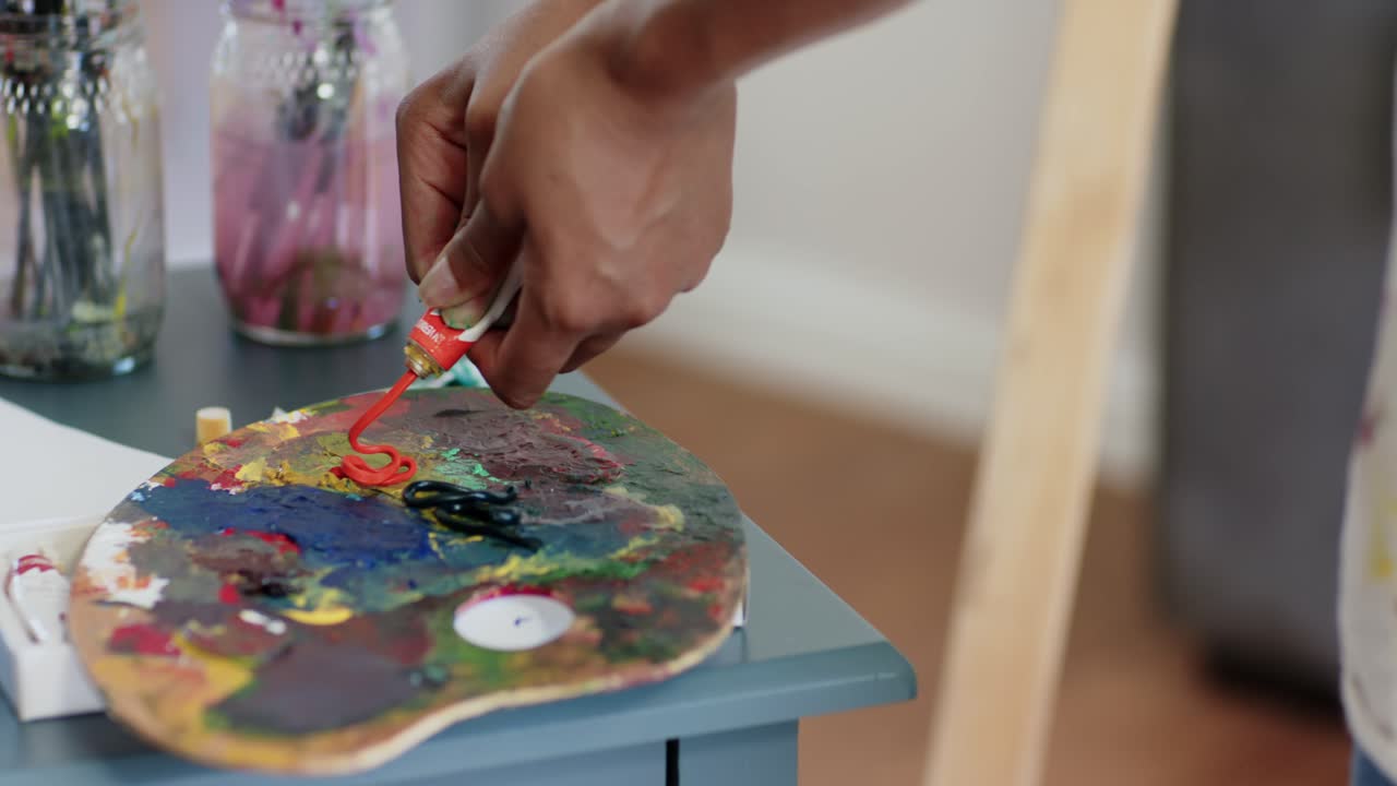 Mulher africana segurando um tubo, espremendo tinta vermelha em espiral na paleta no estúdio, preparando a tinta.
