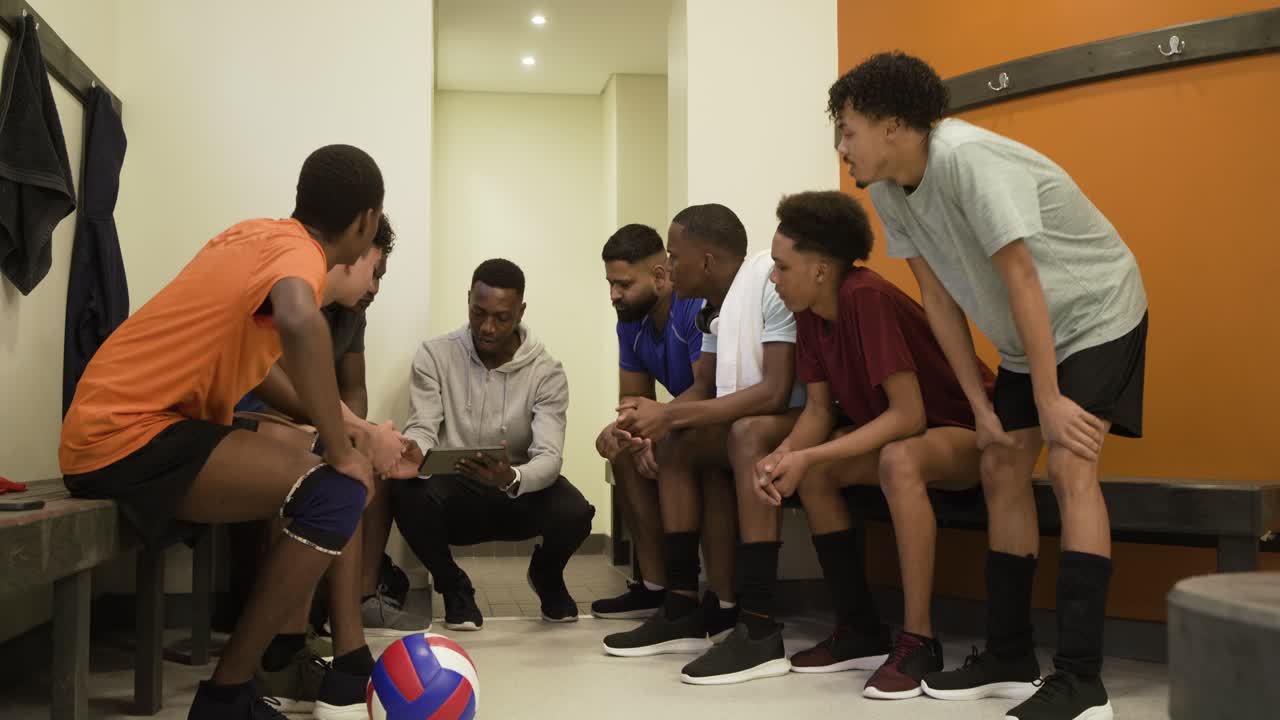 Ein afroamerikanischer Trainer kniet in der Umkleidekabine, zeigt auf ein Klemmbrett und fordert die Männer auf, den Spielzug zu studieren.