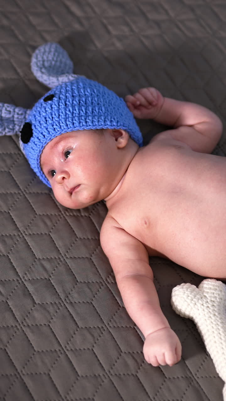 Cute baby in a funny blue suit lying on the bed. Puppy costume put on a newborn kid and funny soft knitted bone beside. View from above. Vertical video