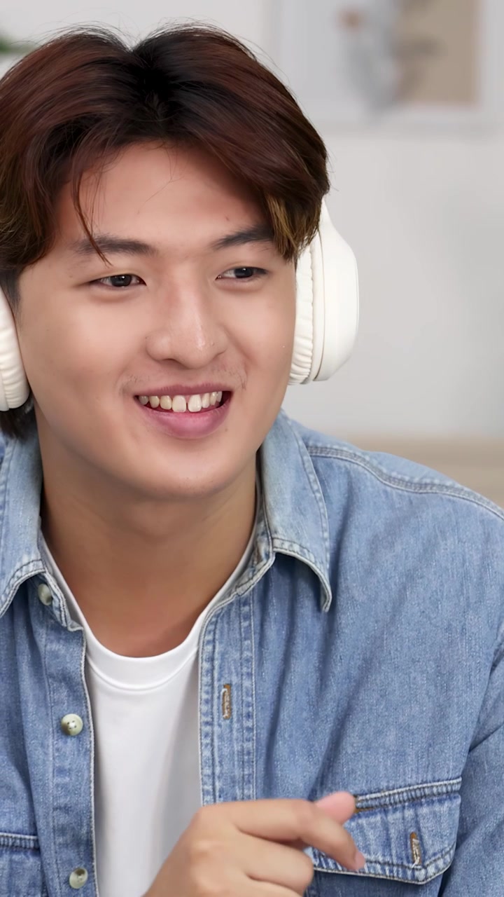 Un giovane uomo asiatico indossa cuffie bianche e una camicia di jeans mentre sorride al chiuso