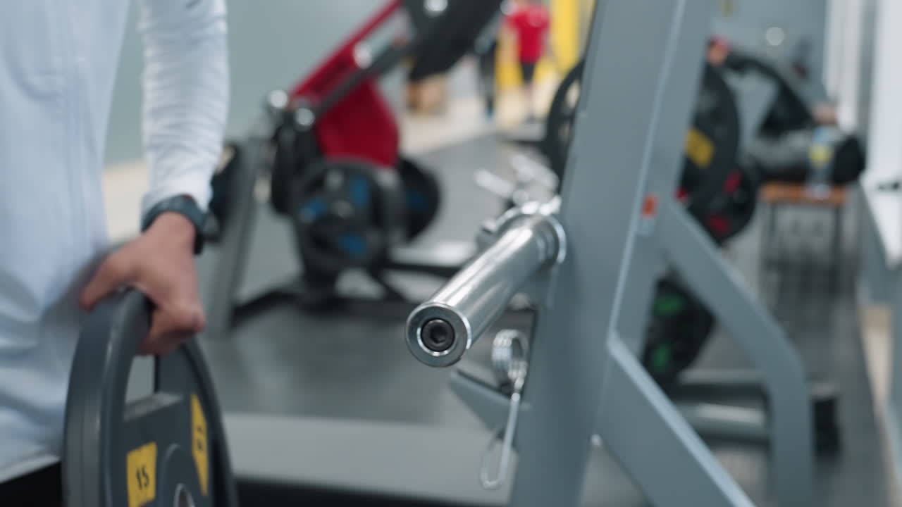Close up of man removing weight plate from barbell sleeve with blurred view of gym members working out, showing hands guiding heavy metal plate off rack during training equipment adjustment session