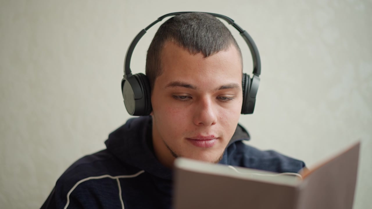 Close up of young man wearing headphones reading book indoors soft lighting calm expression focused on text plain background closeup indoor headset focusing