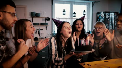 chica europea feliz sostiene pastel de cumpleaños, sopla en velas celebrando con amigos multiétnicos y confeti cámara lenta