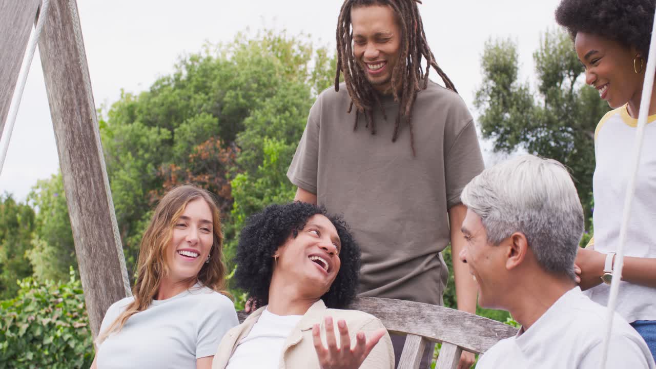 Vertical video: Diverse friends on swing bench in garden, center man telling story evoking laughter