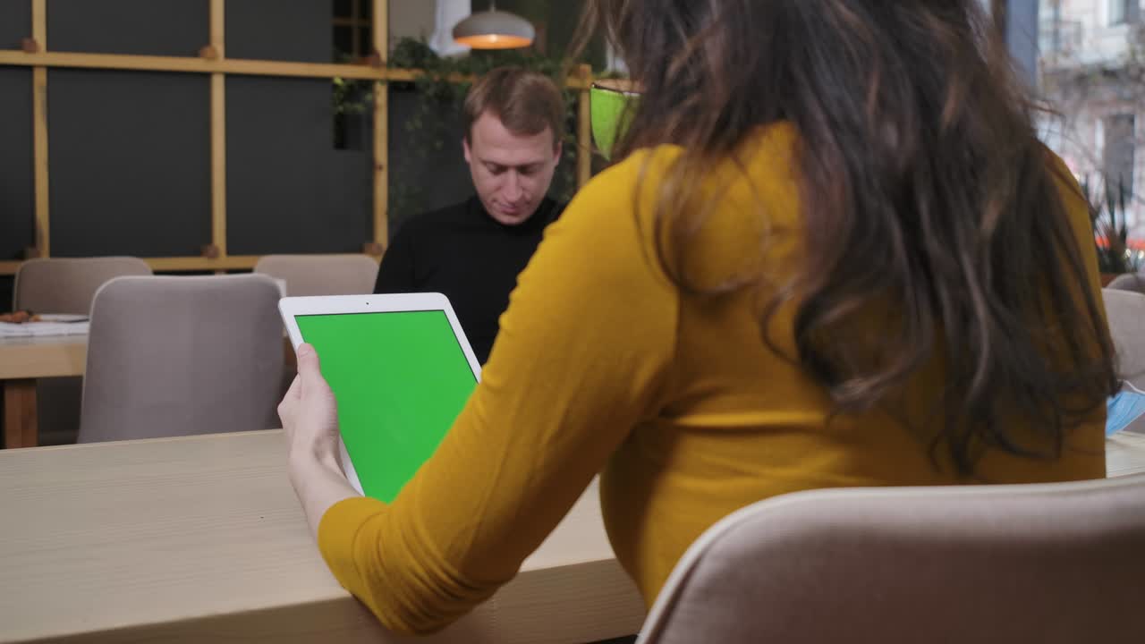 disparando por encima del hombro de una joven y delgada mujer caucásica sentada en un restaurante usando una tableta con pantalla verde y un hombre borroso navegando por internet en un teléfono inteligente en el fondo. cine 4k prores hq.