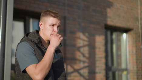 portrait view of young boy standing beside brick building raises cigarette to lips with hesitation before discarding it into nearby waste bin next to wooden bench under bright sunlight
