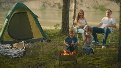 Two-boys-3-6-years-together-fry-on-fire-marshmallows-on-sticks-against-the-background-of-parents.-Family-hike-in-the-woods-with-a-tent.-Family-in-nature-on-a-hike