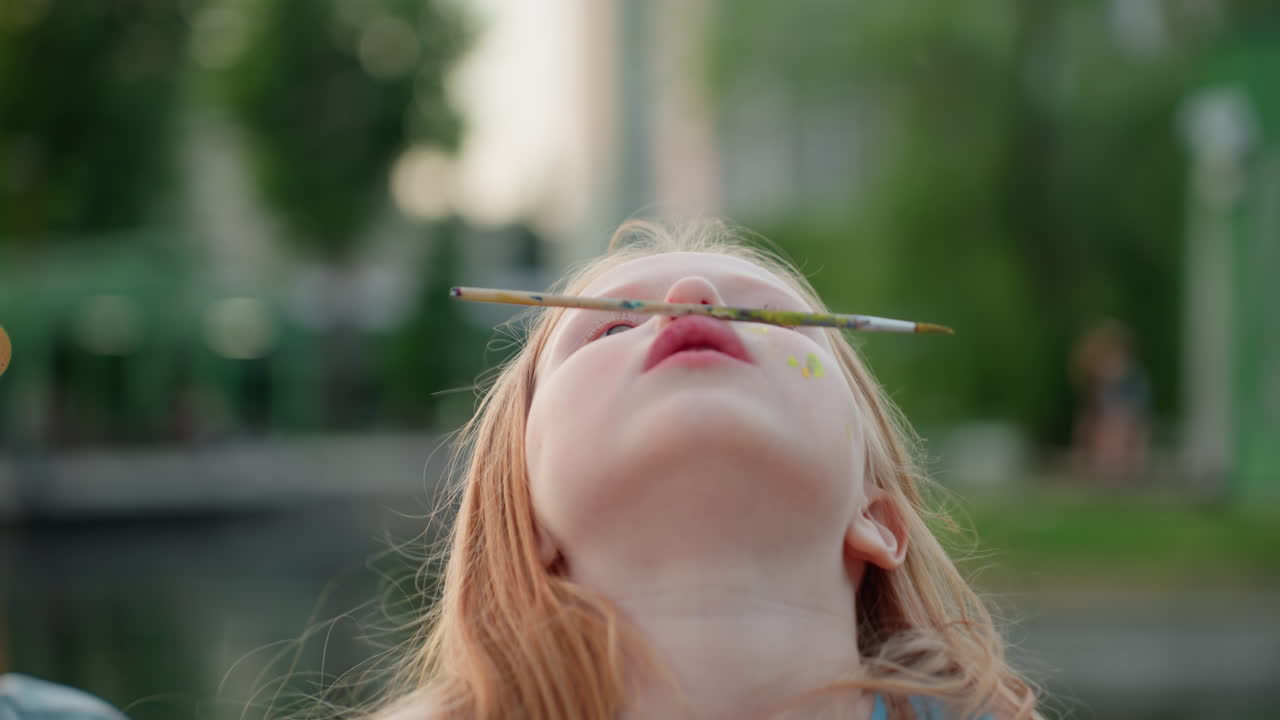 close up of girl looking up balancing pencil on nose with playful expression at golden hour park, blurred adult figure behind, creative moment with bright paint dots on cheek, whimsical art scene