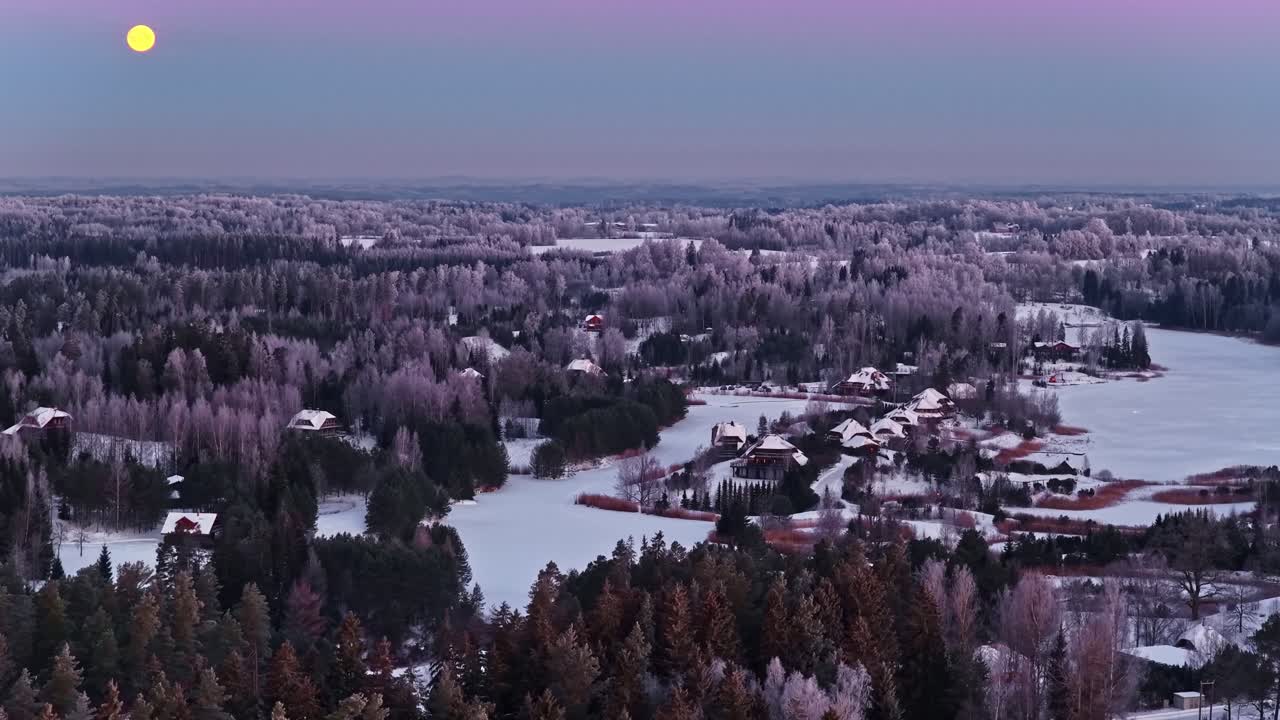 Tasainen ilmakuva lumisen metsän ja maalaiskylän yllä pehmeän pastellisävytteisen iltahämärän taivaan alla