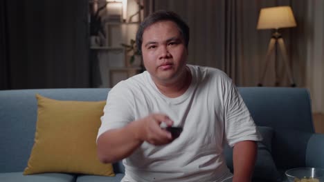 asian fat male with tv remote pressing button and changing, switch channels while sitting on a couch at home