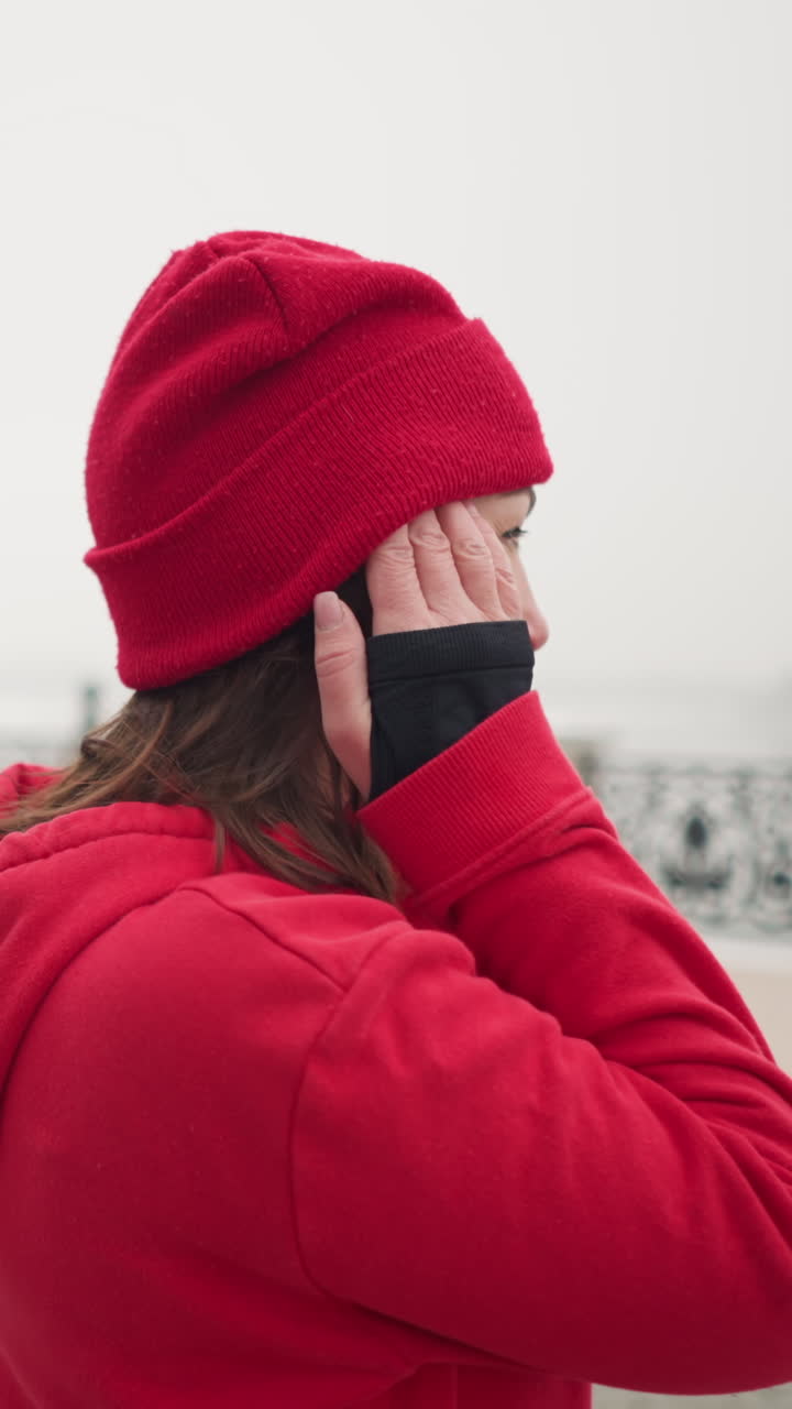 vista trasera de una mujer con capucha roja ajustando su gorra roja al aire libre durante el invierno, entorno urbano con alrededores cubiertos de nieve y barandillas decorativas visibles en el fondo bajo clima de niebla
