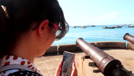 mujer filmando con el teléfono algunos viejos cañones frente a algunos barcos navegando en el océano