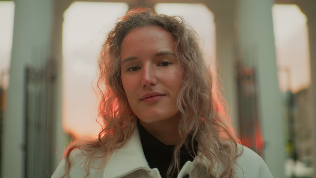 portrait view of woman posing with calm smile outdoors during sunset standing in front of white pillars with warm lighting on face and hair, expressing relaxed mood in urban setting