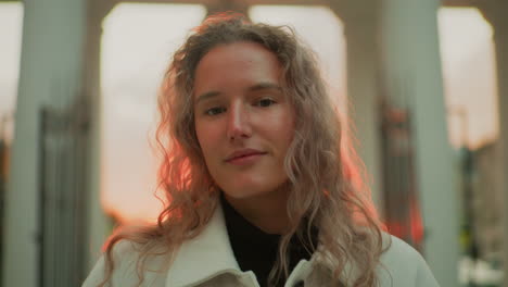 portrait view of woman posing with calm smile outdoors during sunset standing in front of white pillars with warm lighting on face and hair, expressing relaxed mood in urban setting