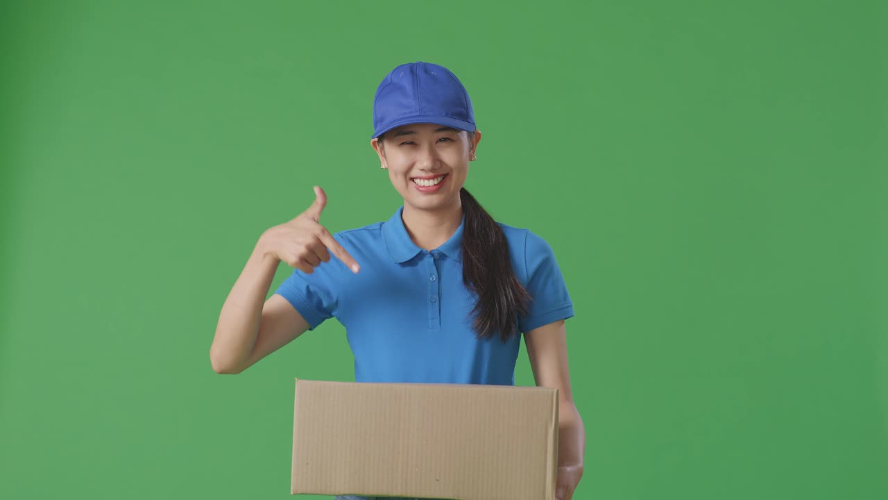 Asian Female Courier In Blue Uniform Smiling And Pointing To A Carton In Hand While Delivering It On Green Screen Background In The Studio