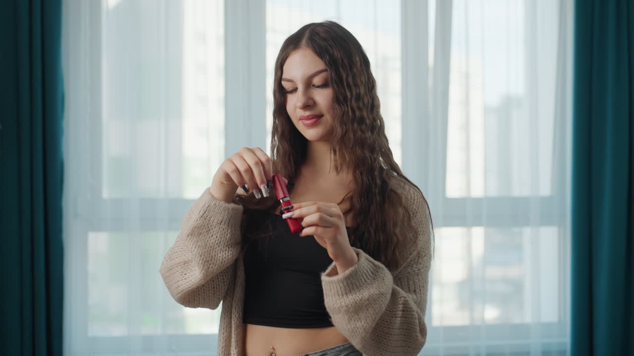 Caucasian Woman Applying Lipstick By Window, Beauty Influencer Testing Red Shade During Morning Routine In Apartment, Curly Brunette Hair, Cozy Cardigan And Black Crop Top, Soft Natural Light And City