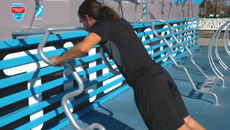 hispanic man with ponytail does incline push ups on outdoor fitness course push up bar, medium shot