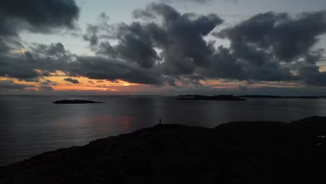 Drohnenaufnahme-Einer-Personensilhouette-Am-Rande-Eines-Berges-Mit-Meer-Im-Hintergrund-Zur-Goldenen-Stunde-Mit-Dramatischer-Wolkenlandschaft