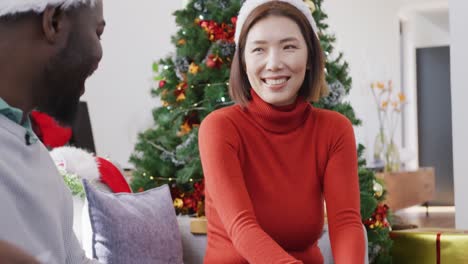 video of happy diverse couple in santa hats exchanging christmas gift and smiling at home