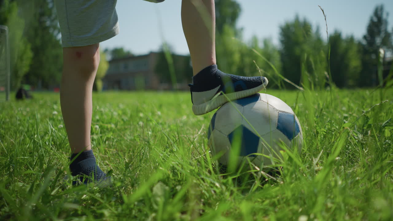 vista de cerca de la pierna de un niño pequeño con un pie en una pelota de fútbol en un campo cubierto de hierba, el fondo presenta vistas borrosas de árboles, un edificio y una carretera pavimentada bajo un cielo soleado