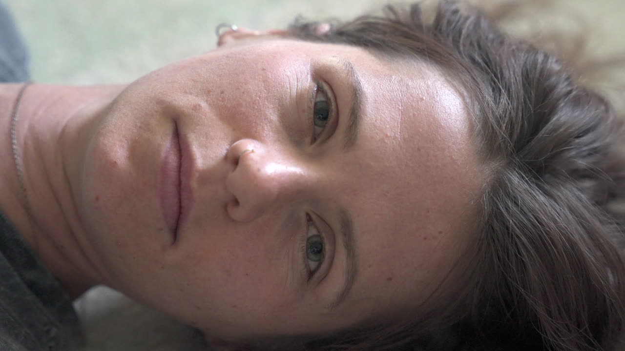 A close-up shot of a woman lying on the ground, opening her eyes and gazing into the camera. The camera slowly rotates around her, capturing her expression.