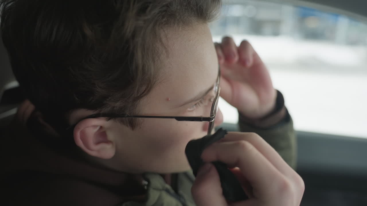 Young boy cleaning glasses lens with soft cloth inside car interior blur building visible through window under soft natural light and subtle reflections highlighting careful hand motion detail